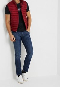 Röd pufferväst över en svart t-shirt, kombinerad med blå jeans och vita sneakers. Har en figurnära design och slät textur.