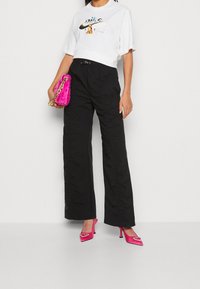 Kvinna klädd i vit grafisk Nike T-shirt, svarta högmidjade vida byxor, klarröda klackar med hjärtdekor och bär en rosa kedjeväska.