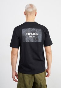Camiseta de algodón negra con un gran gráfico gris que presenta la palabra "DICKIES" y "SINCE 1922" en la parte trasera, combinada con pantalones cargo verdes.