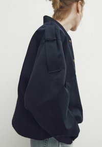 Veste bleu marine en coton, présentant une coupe carrée. Elle possède des épaules marquées, un col montant et des boutons dorés.