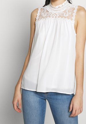 Femme portant un chemisier blanc sans manches avec un détail en dentelle sur le plastron, associé à un jean bleu sur fond uni.