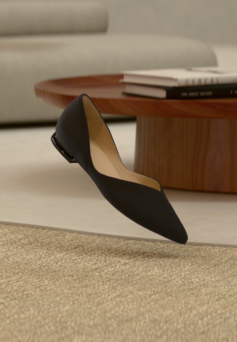 Chaussure plate noire à bout pointu suspendue au-dessus d'un tapis beige texturé, avec une table basse en bois et des livres empilés flous en arrière-plan.