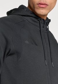 Μαύρο φούτερ με φερμουάρ, με υφασμάτινη υφή στο πάνω μέρος, ρυθμιζόμενο κορδόνι και διακριτικό λογότυπο τριών  ρίγων στο στήθος. Ομαλή υφή υφάσματος.