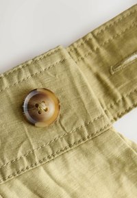 Gros plan sur un bouton marron cousu sur un tissu beige avec des points de couture visibles et une boutonnière partiellement visible.