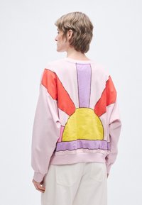 Ružová mikina s farebným dizajnom slnka na chrbte, s oranžovými, žltými a fialovými akcentmi. Pohodlný strih s rebrovaným výstrihom.