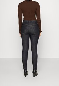 Kvinna klädd i mörkblå skinny jeans, brun ribbad höghalsad tröja och svarta högklackade ankelstövlar, vänd bort från kameran mot en enfärgad bakgrund.