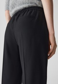 Pantalon noir en tissu texturé, avec une taille élastique et des poches latérales, montré de l'angle arrière. Haut gris clair partiellement visible.