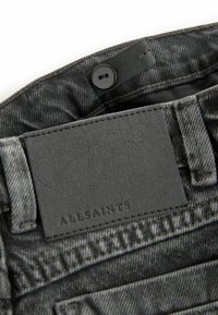 Svart denimtyg med en strukturerad yta, med en grå läderlapp märkt "ALLSAINTS." Inkluderar en svart knappstängning.