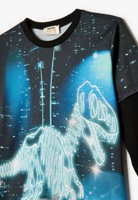 Sort langærmet t-shirt med et lysende dinosaur-skelet grafik mod en blå og sort bylandskab baggrund. Blødt stof, rund halsudskæring.
