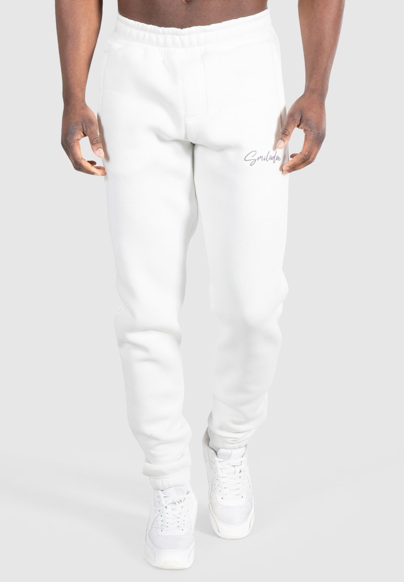 Weiße Baumwoll-Sweatpants mit einem elastischen Bund, schmal zulaufenden Beinen und einem kleinen gestickten Logo auf der linken Seite. Getragen mit weißen Turnschuhen.