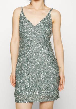 Femme portant une mini robe sans manches en sequins argentés avec fines bretelles et décolleté en V, debout devant un fond blanc uni.