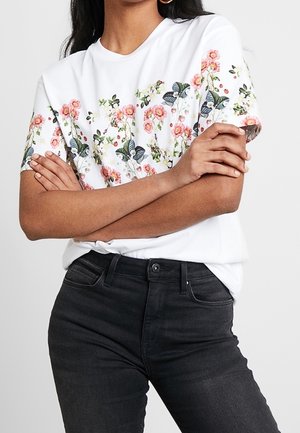 Biała koszulka z kwiatowym wzorem różowych kwiatów i zielonych liści. Połączona z czarnymi dżinsami, prezentująca swobodny strój.