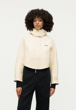 Kobieta o kręconych włosach, ubrana w beżową kurtkę z wysokim kołnierzem i czarne spodnie, stoi na jednolitym białym tle.