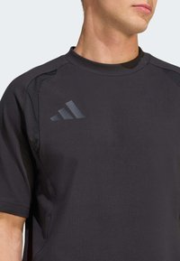 Camiseta deportiva negra de manga corta, con cuello redondo y detalles laterales. El logo de Adidas aparece en un acabado mate sutil en la parte delantera.