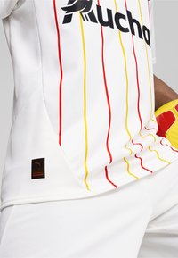 Camiseta de fútbol blanca con franjas verticales rojas y amarillas, con un logo negro y una etiqueta que indica "FÚTBOL AUTÉNTICO". Tejido texturizado.