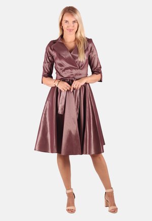 Blonde Frau, die ein glänzendes, mauvefarbenes Wickelkleid mit dreiviertellangen Ärmeln und beigefarbenen Knöchelriemen-Absätzen trägt, stehend vor weißem Hintergrund.