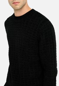 Maglione nero con texture, motivo a griglia, collo a giro e maniche lunghe. Il materiale appare morbido e spesso, offrendo calore e comfort.