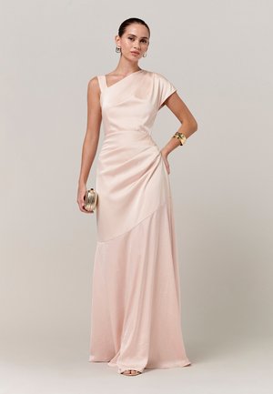 Žena u svijetloružičastoj satenskoj haljini s jednim ramenom, drži zlatnu torbicu, ukrašena zlatnim naušnicama i narukvicama, stoji na sivoj pozadini.