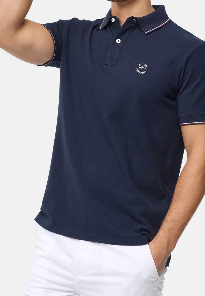 Polo shirt bleu marine avec un col classique, des manches courtes et des accents à rayures blanches et rouges. Présente un logo rond sur la poitrine.