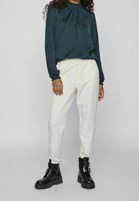 Długa bluzka z ciemnozielonego satynu z długimi rękawami i wysokim dekoltem, zestawiona z kremowymi, zwężanymi spodniami oraz czarnymi botkami na kostkę z ozdobną klamrą.