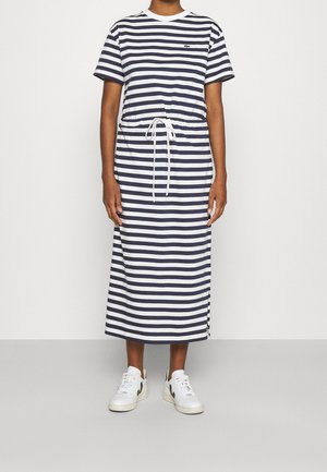 Personne portant une robe midi rayée bleu marine et blanc à manches courtes, ceinture à la taille et baskets blanches, debout sur un sol gris.
