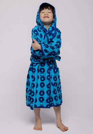 Enfant souriant avec les bras croisés, portant un peignoir à capuche bleu orné de motifs de pommes bleu foncé, debout pieds nus sur un fond clair.
