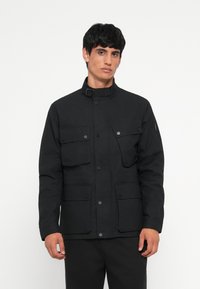 Black utility jacket with a high collar, multiple front pockets, snap fasteners, and a matte finish. Straight cut with long sleeves.