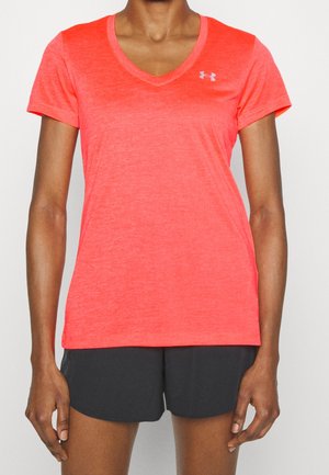 Personne portant un t-shirt de sport corail vif à manches courtes et col en V et un short noir, debout avec les bras détendus le long du corps.
