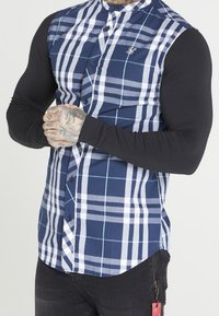 Camicia button-up con un design a quadri blu navy e bianco a maniche corte, abbinata a maniche lunghe nere e un piccolo logo decorativo.