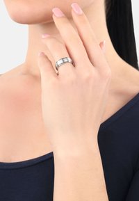 Una mujer se toca la cara con una mano que muestra un anillo de plata, contra un fondo neutro, vistiendo una blusa oscura.