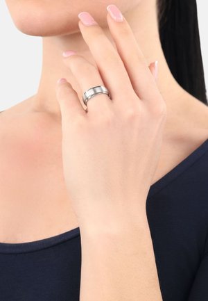 Eine Frau berührt ihr Gesicht mit einer Hand, die einen silbernen Ring trägt, vor einem neutralen Hintergrund, gekleidet in einem dunklen Oberteil.