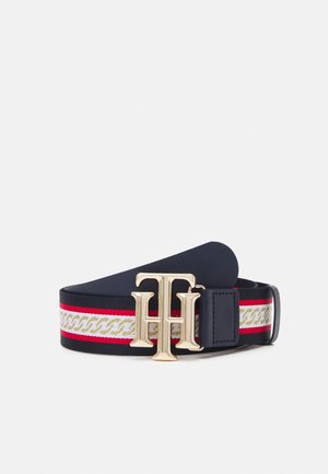 Marineblauwe riem met rood-wit-gouden kettingpatroon, gouden "TH" gesp en zwarte leren accenten, opgerold op een witte achtergrond.