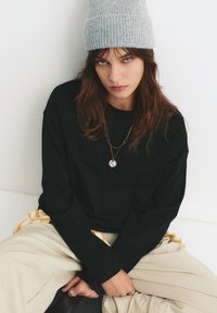 Zwarte lange mouwen shirt, beige broek, grijs gebreide beanie. Een ketting met een ronde hanger, losse stijl, natuurlijke stoffen texturen.