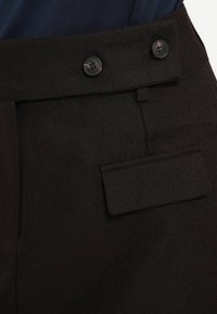 Pantalones negros de tela texturizada, con dos grandes botones en la cintura y un bolsillo trasero con tapa.
