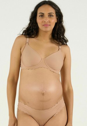 Femme enceinte aux longs cheveux foncés portant un soutien-gorge et des sous-vêtements de maternité beige, debout devant un fond clair uni.