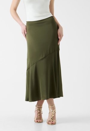 Femme portant une jupe midi asymétrique vert olive, un top blanc sans manches et des sandales à talons hauts dorées à brides sur un fond blanc.