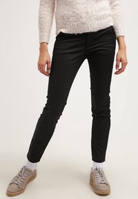 Pantalons noirs slim en coton extensible, avec une texture douce et un design à cinq poches, associés à des baskets grises et des chaussettes blanches.