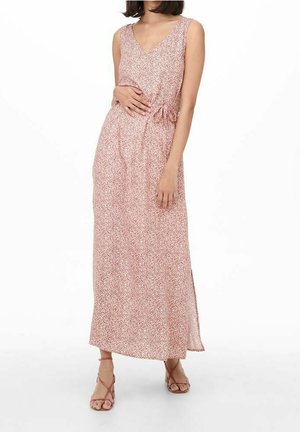 Femme portant une robe longue sans manches à fleurs roses et blanches avec un col en V et une taille nouée, associée à des sandales beiges à brides.