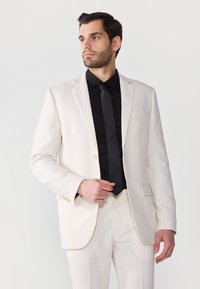 Hellbeige Leinenanzugjacke und -hose, kombiniert mit einem schwarzen Hemd und einer Krawatte. Verfügt über einen Zwei-Knopf-Verschluss und Taschenverzierungen.