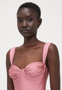 Abito rosa con dettaglio arricciato sul busto, spalline larghe, texture liscia, silhouette aderente e una leggera luminosità.