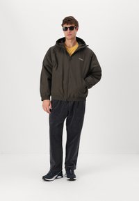 Veste zippée vert foncé avec capuche, associée à un t-shirt jaune et un pantalon en velours côtelé gris foncé. Des baskets noires et des lunettes de soleil complètent le look.