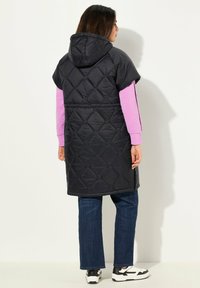 Černá prošívaná vesta s kapucí, krátkými rukávy a dlouhým střihem. Nositá přes růžovou dlouhou košili, spolu s modrými džíny a černobílými teniskami.