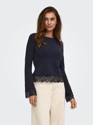 Femme portant un haut bleu marine à manches longues avec bordure en dentelle aux manches et à l'ourlet, associé à un pantalon large beige clair, debout devant un fond uni.