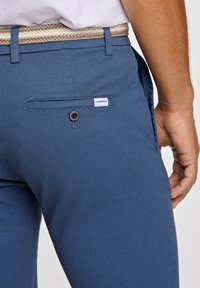 Pantaloni blu texture con tasca posteriore, dotati di un bottone nero e un'etichetta bianca. Cinturati con una cintura intrecciata color tan.