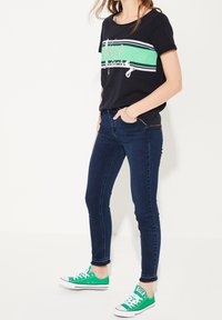 Sort T-shirt med grønne og hvide vandrette striber og et logo, parret med mørkeblå skinny jeans og grønne sneakers. Enkel casual stil.