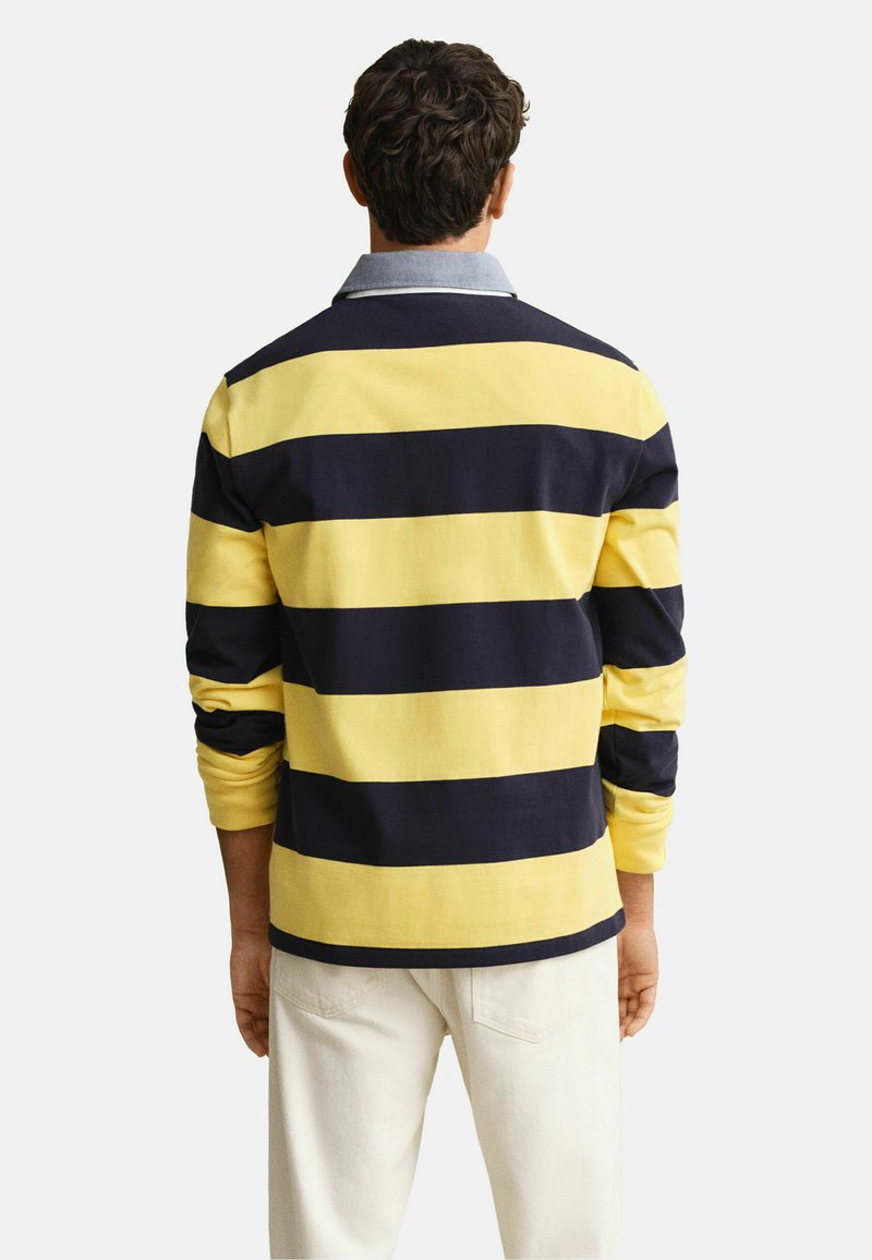 Mann, von hinten gesehen, trägt ein langärmeliges Rugbyhemd mit breiten gelben und marineblauen Horizontalstreifen und hellen Hosen.