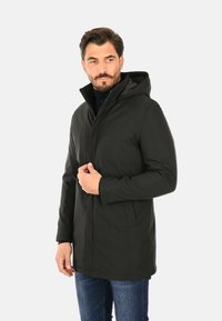 Parka nera impermeabile con cappuccio, dotata di zip frontale, tasche laterali e una texture liscia. Indossata sopra un maglione blu navy e jeans.