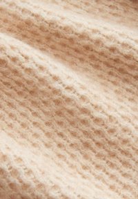 Tessuto beige chiaro con un motivo a waffle testurizzato. Superficie morbida e pelosa, che mette in risalto il calore e la qualità tattile del materiale.