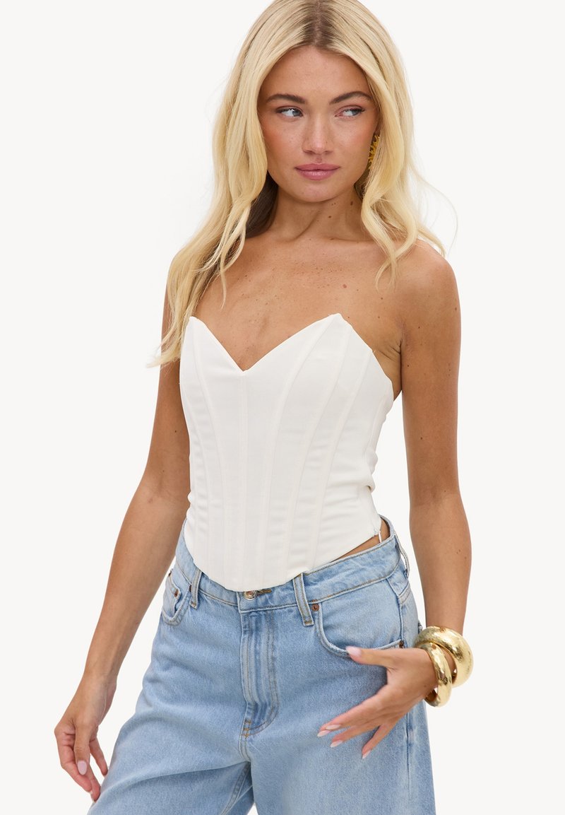 Femme aux longs cheveux blonds porte un corset blanc sans bretelles, un jean bleu clair et des bracelets dorés, regardant vers sa gauche.