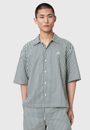 Mann trägt kurzärmliges Hemd mit Knöpfen und passende Hose mit vertikalen schwarz-weißen Streifen, steht mit einer Hand in der Tasche.